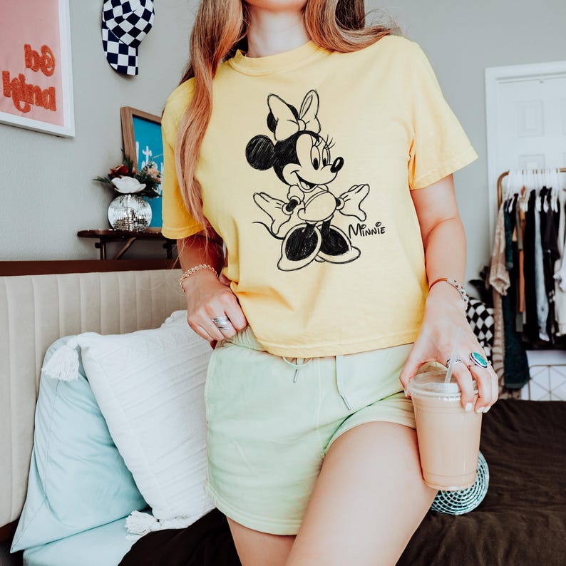 May include: Yellow t-shirt featuring a black and white Minnie Mouse graphic. The shirt has short sleeves and a crew neck. The word "Minnie" is written in cursive below the character. The model is wearing light green shorts and holding a beverage.