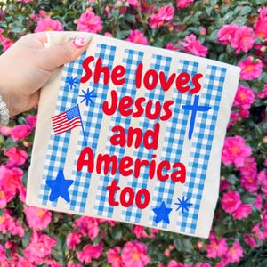 May include: A square, off-white fabric item with a blue and white gingham pattern. The text "She loves Jesus and America too" is written in red. The design includes a blue cross, an American flag, and blue stars.