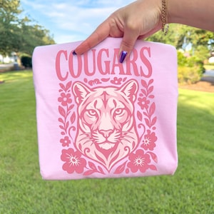 May include: A light pink t-shirt featuring a graphic print of a cougar head surrounded by floral elements. The word COUGARS is printed in a bold, stylized font above the cougar illustration. The design uses various shades of pink and magenta.