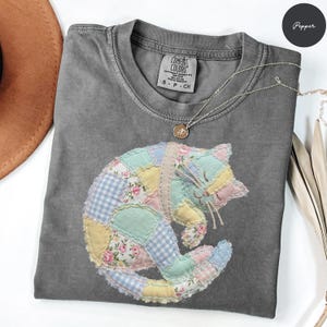 May include: A gray Comfort Colors t-shirt with a patchwork cat design in pastel colors. The cat is made of various fabrics, including floral and gingham patterns. A silver necklace with a small pendant is also visible.