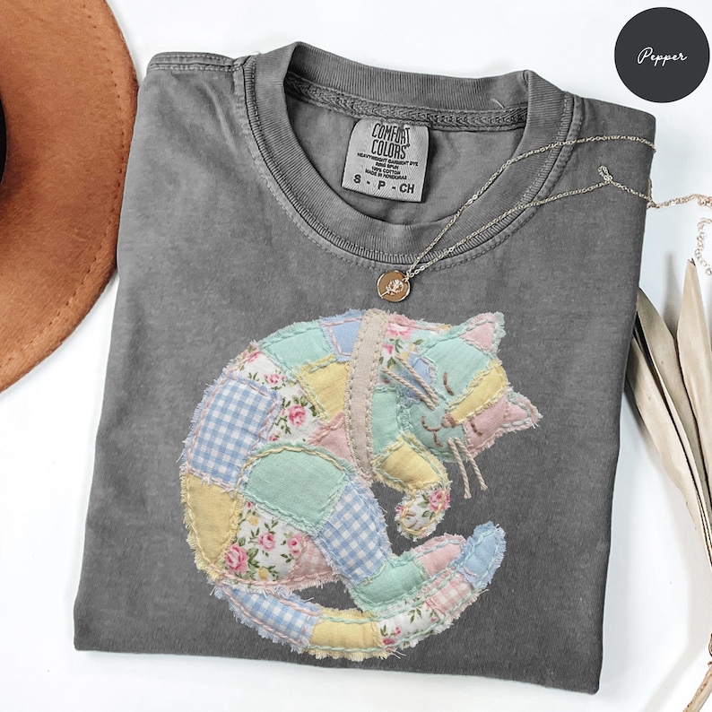 May include: A gray Comfort Colors t-shirt with a patchwork cat design in pastel colors. The cat is made of various fabrics, including floral and gingham patterns. A silver necklace with a small pendant is also visible.