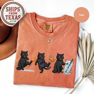 Puede incluir: Camiseta color &ntilde;ame con cuello redondo y un gr&aacute;fico de tres gatos negros. Un gato patea una pelota, otro lleva una corona y el tercero sostiene una bebida. La camiseta tiene un lavado vintage y una etiqueta Comfort Colors.