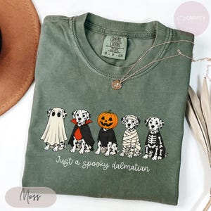 Może przedstawiać: Koszulka w kolorze mchu z grafiką przedstawiającą pięć dalmatyńczyków w kostiumach na Halloween, w tym ducha, wampira, dynię, mumię i szkielet. Poniżej psów znajduje się napis Just a spooky dalmatian wykonany białą, ozdobną czcionką.