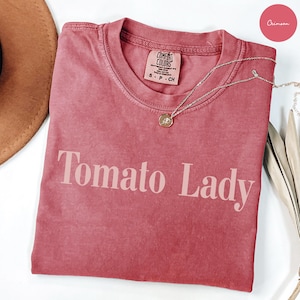 May include: A crimson short-sleeved t-shirt with a crew neckline and the text Tomato Lady printed in a light pink font across the chest. The shirt features a Comfort Colors brand label at the inner neck and is paired with a thin gold-toned necklace.