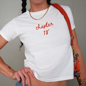 Peut inclure: T-shirt blanc avec le texte rouge "chapter 18". Le t-shirt est associé à un collier chaîne doré et un sac à bandoulière orange. L'image montre une personne portant le t-shirt.