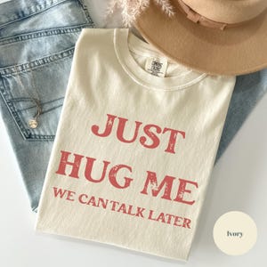 May include: Ivory-colored t-shirt with the text "JUST HUG ME WE CAN TALK LATER" in red. The shirt is folded on top of blue jeans, with a gold necklace and a tan hat visible. The shirt is a Comfort Colors brand.