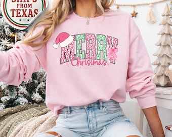 May include: A light pink sweatshirt with the words "MERRY Christmas" in a green and pink leopard print design. The word "MERRY" has a pink Santa hat on top. The sweatshirt is a long-sleeved crew neck.