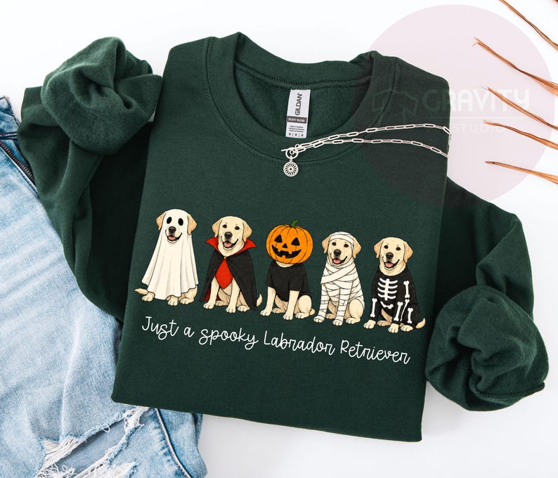Może przedstawiać: Ciemnozielona bluza z okrągłym dekoltem z grafiką przedstawiającą pięć labrador&oacute;w w kostiumach na Halloween, w tym ducha, wampira, dynię, mumię i szkielet. Tekst pod psami brzmi: "Just a spooky Labrador Retriever."