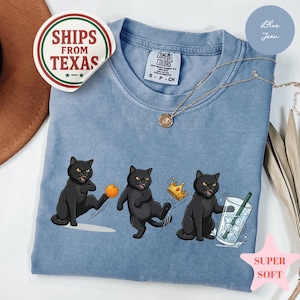 Puede incluir: Camiseta azul vaquero claro con un gr&aacute;fico de tres gatos negros jugando con una pelota, con una corona y sosteniendo una bebida. La camiseta tiene el texto "COMFORT COLORS" y "SUPER SOFT". Un collar est&aacute; drapeado sobre la camiseta.