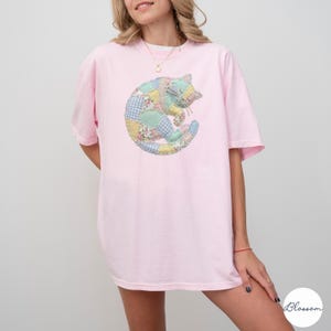 May include: A light pink t-shirt featuring a colorful patchwork cat design. The cat is curled up, with a mix of floral, gingham, and solid fabric patches. The shirt has short sleeves and a relaxed fit.