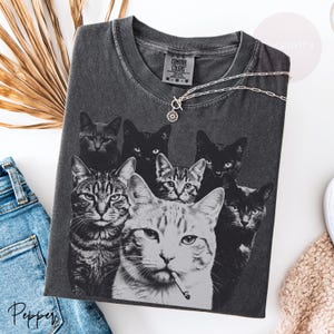 May include: A charcoal gray t-shirt featuring a graphic print of seven cats in various poses, including one cat with a cigarette in its mouth. The shirt has a Comfort Colors brand label at the neck and a silver-toned chain necklace resting on top.