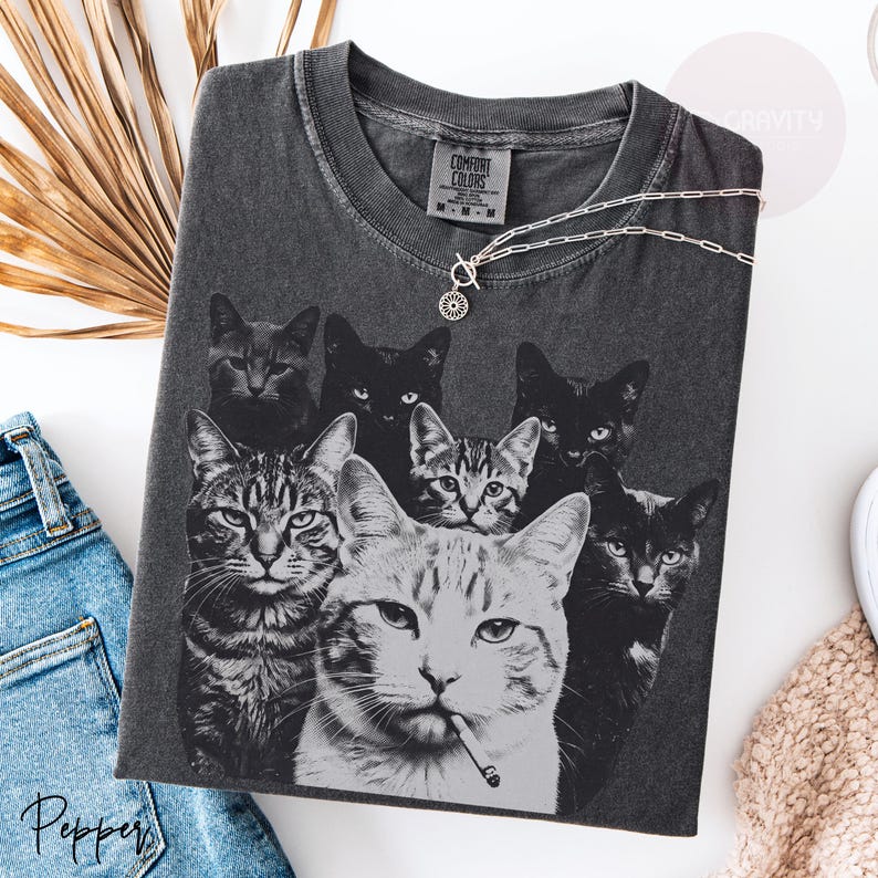 May include: A charcoal gray t-shirt featuring a graphic print of seven cats in various poses, including one cat with a cigarette in its mouth. The shirt has a Comfort Colors brand label at the neck and a silver-toned chain necklace resting on top.