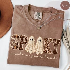 May include: An espresso-coloured t-shirt featuring the word SPOOKY with two ghost-shaped letters in the centre. The letters have various patterns including plaid, floral, and leopard print. The text custom your text is printed below.