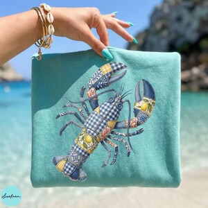 May include: A turquoise zippered pouch featuring a patchwork lobster design. The lobster is detailed with blue gingham, floral, and lemon patterns. The pouch is held up by a hand with a gold bracelet. The word "Seafoam" is printed on the bottom left.