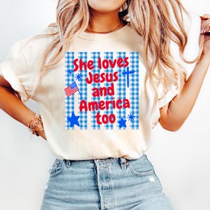 May include: Cream-colored t-shirt with the text "She loves Jesus and America too" in red and blue. The design includes an American flag, a cross, and blue stars on a blue and white checkered background.