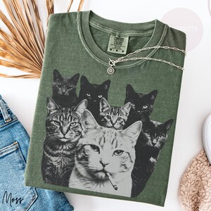 May include: A moss green crew neck t-shirt featuring a graphic print of seven cats in various poses. The central cat is depicted with a cigarette in its mouth. The shirt has a Comfort Colors label at the inner neckline and a short-sleeved design.
