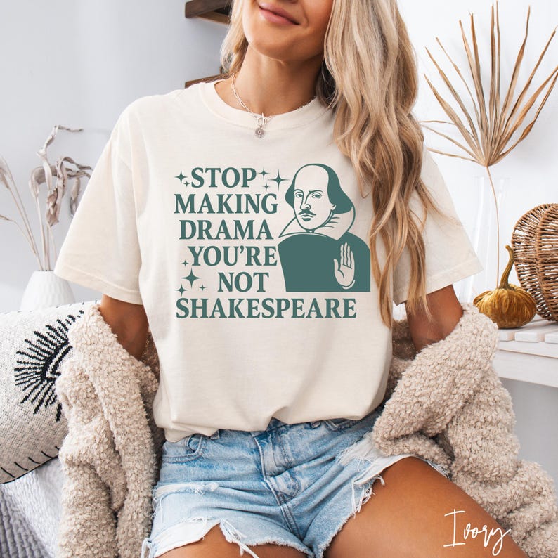 May include: An ivory short-sleeve t-shirt featuring a dark green graphic of William Shakespeare with a hand raised in a stop gesture. The text reads: STOP MAKING DRAMA YOU'RE NOT SHAKESPEARE, accented with small four-pointed star icons.