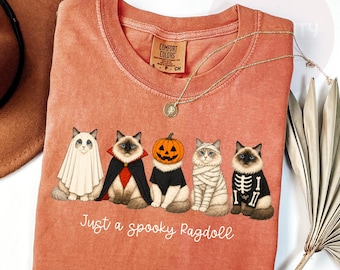May include: A terracotta-colored crewneck t-shirt featuring a graphic of five Ragdoll cats in Halloween costumes, including a ghost, vampire, pumpkin, mummy, and skeleton. The text below the cats reads, "Just a spooky Ragdoll".