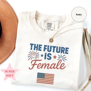 Puede incluir: Camiseta de color marfil con el texto "THE FUTURE IS FEMALE" en azul y rojo, con fuegos artificiales y una bandera americana. La camiseta está etiquetada como "SUPER SOFT" e incluye una etiqueta Comfort Colors.
