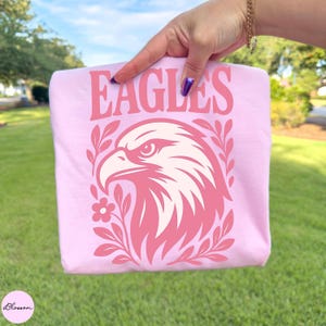 May include: A light pink t-shirt featuring a dark pink graphic of an eagle head in profile, surrounded by stylized leaves and a small flower. The word EAGLES is printed in large, bold, dark pink capital letters above the eagle illustration.