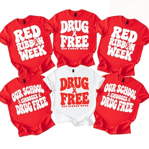 May include: Five short-sleeved t-shirts arranged in a grid. Four are red with white text reading Red Ribbon Week or Our School Chooses Drug Free. One central white t-shirt features red text reading Drug Free Red Ribbon Week with ribbon icons.