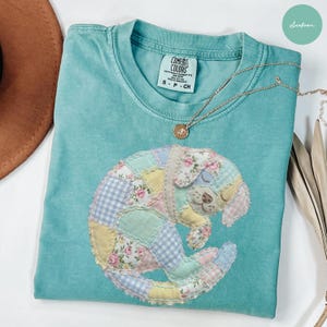 Könnte beinhalten: Türkisgrünes Comfort Colors T-Shirt mit einem Patchwork-Welpen-Design. Der Welpe besteht aus verschiedenen Blumen- und Vichy-Stoffen. Eine goldene Halskette mit Anhänger liegt auf dem Shirt. Rundhalsausschnitt, kurze Ärmel.