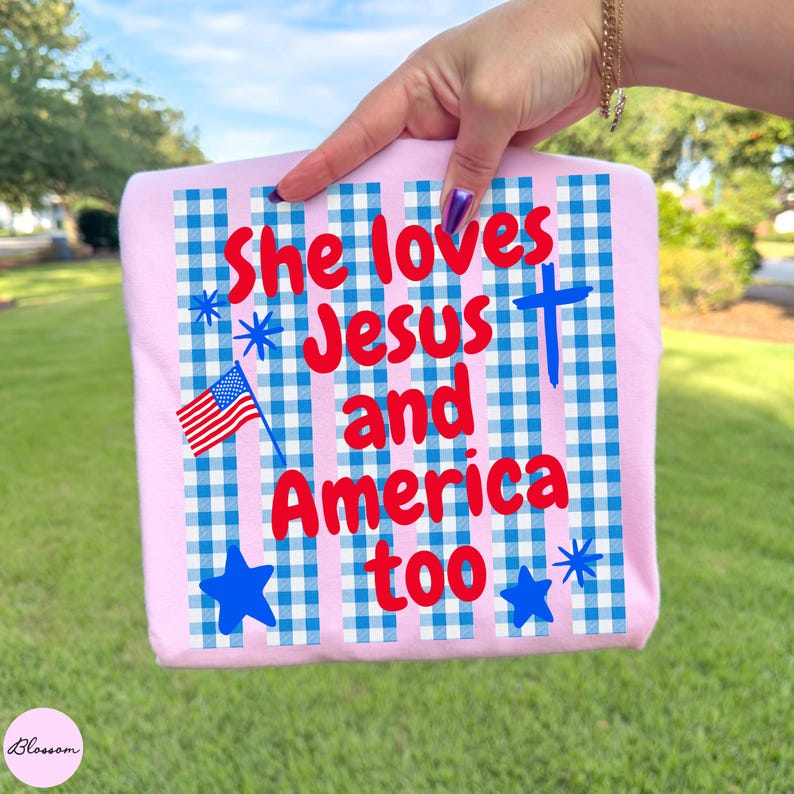 May include: A light pink t-shirt with the text "She loves Jesus and America too" in red. The design includes a blue cross, stars, and a small American flag on a blue and white checkered background.