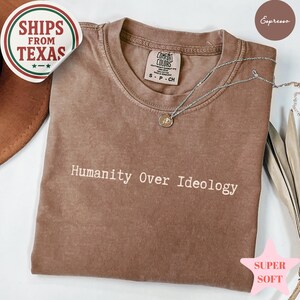 May include: Espresso-colored t-shirt with the text "Humanity Over Ideology" in white. The shirt has a crew neck and short sleeves. A silver necklace with a pendant is draped across the shirt. The shirt is labeled "Super Soft".