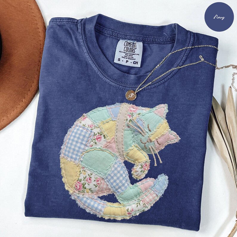 May include: Navy blue t-shirt with a patchwork cat design. The cat is made of various pastel-colored fabrics, including floral and gingham patterns. A gold necklace with a pendant rests on the shirt. The shirt has a Comfort Colors label.