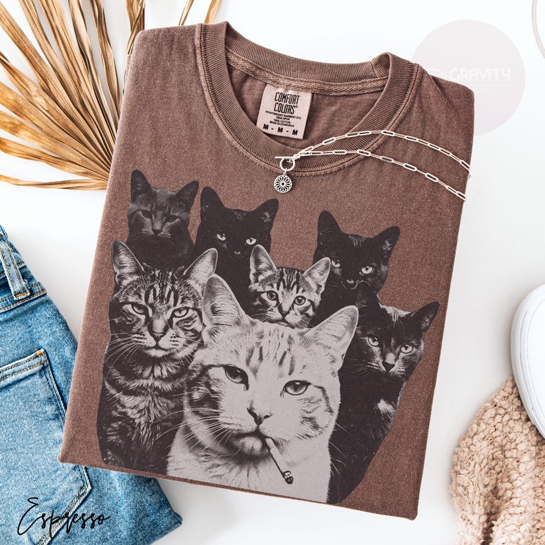 May include: A brown short-sleeve t-shirt featuring a graphic print of six cats, including one tabby cat with a cigarette in its mouth. The shirt has a crew neckline and a Comfort Colors brand label on the inside collar. A silver chain necklace rests on top.