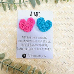 May include: A card with the name "AIMEE" printed on it. Two crocheted hearts, one pink and one blue, are attached to the card. The card reads "A little hug to keep in your bag, for whenever you're feeling a little sad. One for Mummy and one for you, to make us feel better if we're feeling blue." The card also includes the Instagram handle "@stitchedbysarah_scriv".