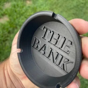 Op de afbeelding: Een zwarte asbak met de tekst "THE BANK" in reliëf op de bodem. De ronde asbak heeft twee inkepingen voor sigaretten. De asbak wordt in een hand gehouden.