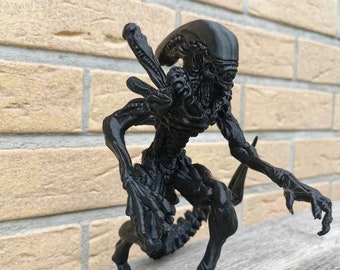 May include: A black 3D printed figurine of an alien creature with a long tail and sharp claws. The figurine is posed in a crouched position, with its head turned to the side.