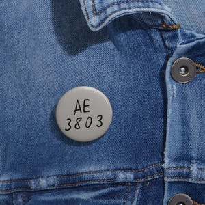 May include: A light gray button pin with the letters "AE" above the numbers "3803" in black. The pin is attached to a blue denim jacket.