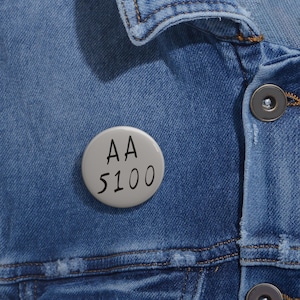 May include: A light gray button pin with the text "AA 5100" in black, attached to a blue denim jacket. The button is round and has a simple, clean design, contrasting with the textured denim.