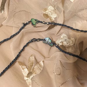 May include: A black cord necklace with two iridescent shell pendants. The necklace is on a beige background with a floral pattern.