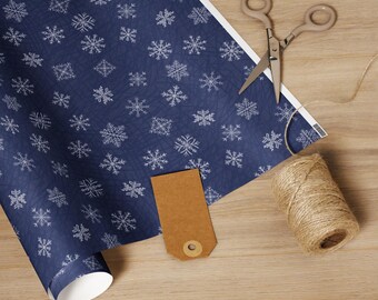 May include: A roll of navy blue wrapping paper with a white snowflake pattern, a pair of scissors, a brown tag, and a spool of twine. The items are arranged on a light brown wooden surface.