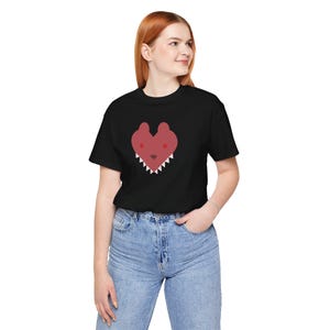 May include: Black t-shirt featuring a red heart-shaped graphic with a bear face design. The bear face has white teeth. The model is wearing blue jeans.
