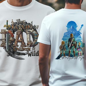 Könnte beinhalten: Weiße T-Shirts mit grafischen Designs. Ein Shirt zeigt den Text "The Legend of Dad" mit einem Zahnrad- und Schwertdesign. Das andere Shirt hat eine farbenfrohe Illustration von Charakteren mit einem Berg im Hintergrund.