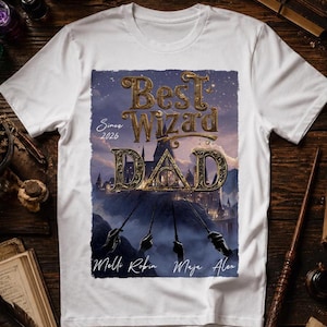 Könnte beinhalten: Weißes T-Shirt mit einem grafischen Design mit den Worten "Best Wizard Dad" in goldfarbenen Buchstaben, mit einer Schlossillustration und den Namen Melli, Robin, Maja und Alex. Das Shirt hat auch den Text "Since 2026".