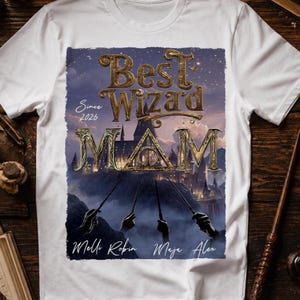 Könnte beinhalten: Weißes T-Shirt mit einem goldenen Aufdruck "Best Wizard Mom" vor einer Schlosskulisse. Das Design enthält den Text "Since 2026" und drei Namen. Das T-Shirt hat kurze Ärmel und ist aus weichem Material.