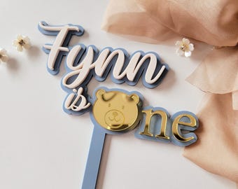 May include: A blue and white cake topper with the name "Fynn" and the words "is one." The topper features a gold bear face and is designed for a first birthday celebration. The letters are layered for a 3D effect.