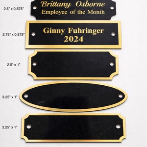 Puede incluir: Una colección de placas y placas de identificación negras y doradas. La placa superior dice "Brittany Osborne, Empleado del mes". La segunda placa dice "Ginny Fuhringer 2024". Se muestran varias formas y tamaños, con medidas en cm.