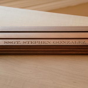 Könnte beinhalten: Ein Namensschild aus Holz mit dem eingravierten Text "SSGT. STEPHEN GONZALEZ" und einem militärischen Emblem. Das Namensschild besteht aus geschichtetem Holz in Brauntönen und hat ein gestuftes Design.