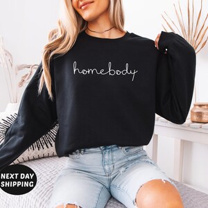 May include: A black sweatshirt with the text "homebody" written in white cursive on the front. The sweatshirt is being worn by a person with long blonde hair and is paired with light blue denim shorts.