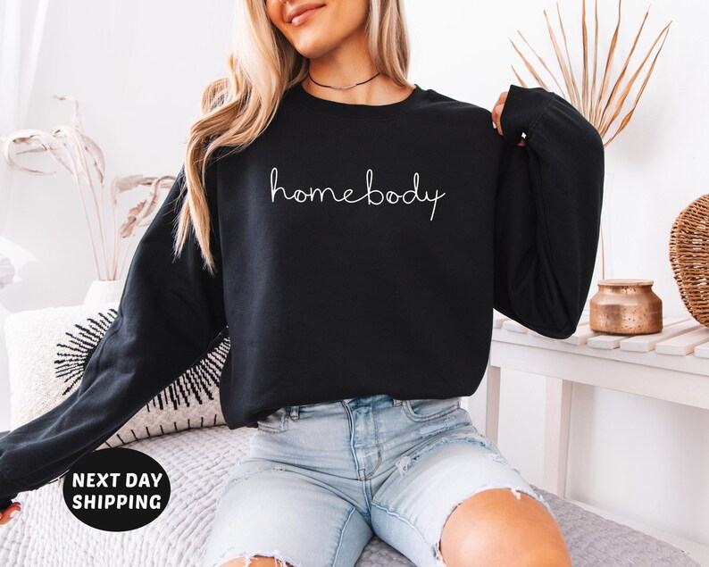 May include: A black sweatshirt with the text "homebody" written in white cursive on the front. The sweatshirt is being worn by a person with long blonde hair and is paired with light blue denim shorts.