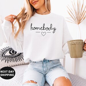 May include: White sweatshirt with the text "homebody" in black script with a small heart underneath.