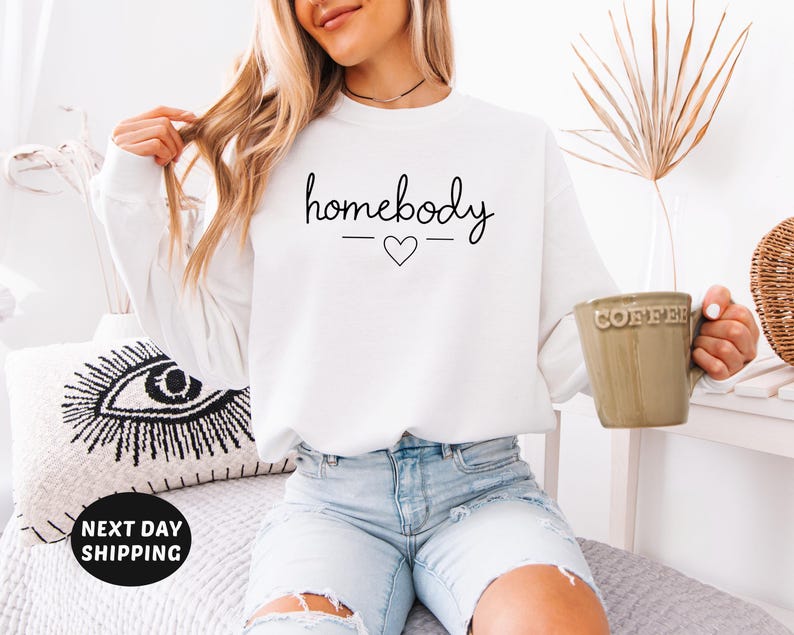 May include: White sweatshirt with the text "homebody" in black script with a small heart underneath.