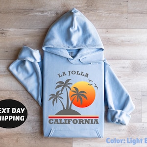 May include: Light blue hoodie with a graphic of palm trees, a sunset, and the text "LA JOLLA CALIFORNIA." The hoodie has a drawstring hood and long sleeves. The color is light blue.