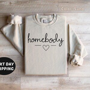 May include: A sand-colored sweatshirt with the text "homebody" in black script with a small heart in between two lines.